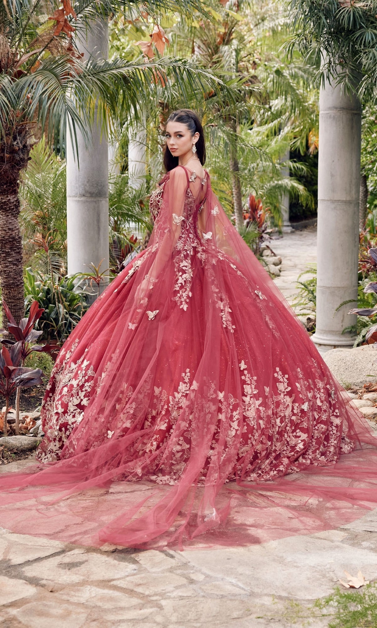 A woman stands outdoors in the Juliet 1442 Quinceañera Dress with Sheer Cape—a voluminous red ball gown featuring gold floral details—surrounded by palm trees and stone pillars.